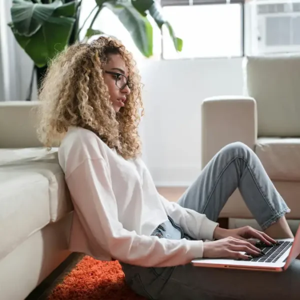 Mulher sentada no chão encostada no sofá usando notebook em ambiente doméstico iluminado. Foto: Thought Catalog/Unsplash