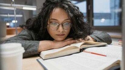 Mulher com óculos e expressão concentrada apoia o rosto nas mãos enquanto lê livros abertos sobre a mesa.