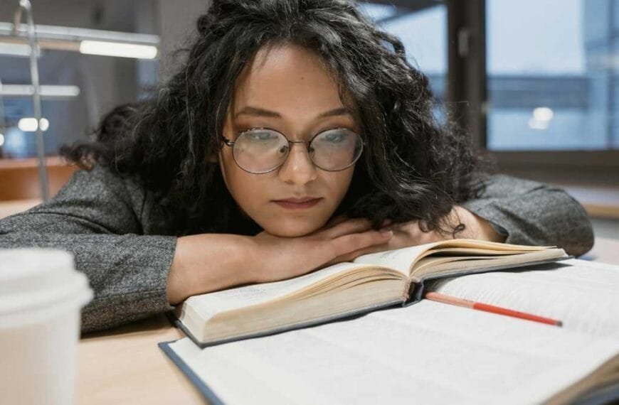 Mulher com óculos e expressão concentrada apoia o rosto nas mãos enquanto lê livros abertos sobre a mesa.