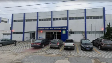 Concursos 7 Fachada do prédio da Câmara Municipal de Mongaguá, com estrutura branca e detalhes em azul, estacionamento frontal com carros e céu nublado ao fundo.