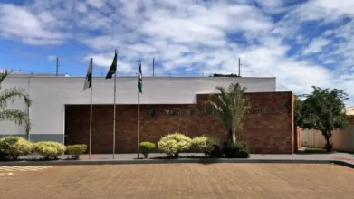 Fachada da Câmara Municipal de Pitangueiras com paredes brancas e revestimento de tijolos, possuindo três mastros com bandeiras à esquerda, vegetação rasteira e céu azul ao fundo.