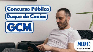 Homem de camiseta branca sentado em um sofá preto, segurando e usando um tablet com uma das mãos, ao lado de uma almofada listrada e uma planta decorativa ao fundo - Concurso Duque de Caxias oferece 500 vagas para GCM