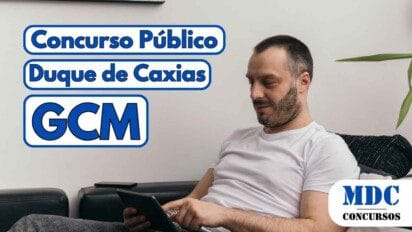 Homem de camiseta branca sentado em um sofá preto, segurando e usando um tablet com uma das mãos, ao lado de uma almofada listrada e uma planta decorativa ao fundo - Concurso Duque de Caxias oferece 500 vagas para GCM