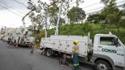 Equipe da CEMIG faz manutenção em rede elétrica urbana com caminhões e cestos aéreos; eletricistas usam EPIs, operam cabos em poste e sinalizam a área com cones.