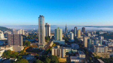Vista aérea da cidade de Francisco Beltrão (PR) com prédios altos, céu azul e neblina ao fundo.