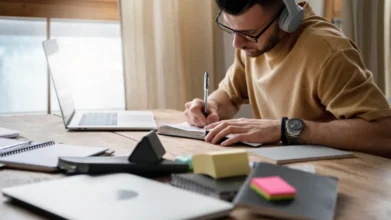 Homem usando fones de ouvido estuda em mesa com notebook, cadernos e post-its, escrevendo em um caderno. Foto: Freepik