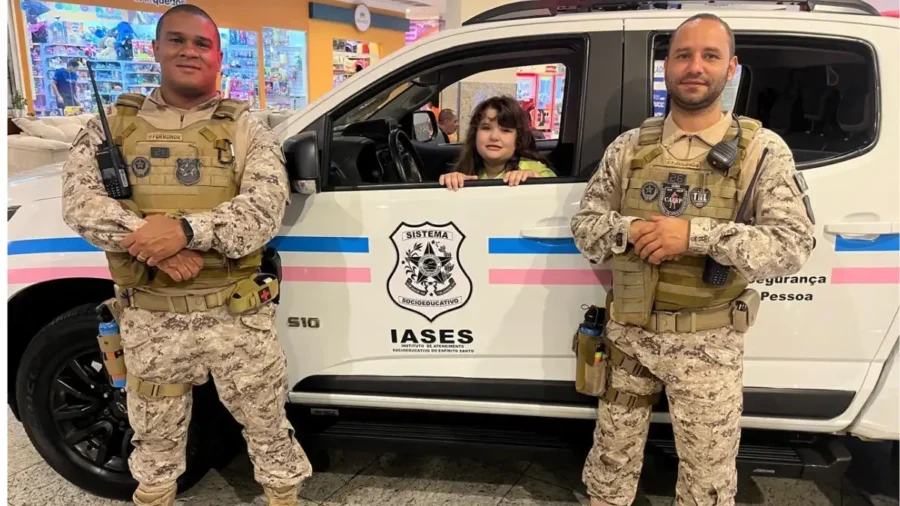 Dois agentes do IASES fardados com colete tático e rádio posam ao lado de viatura S10 com escudo do concurso IASES do Sistema Socioeducativo, enquanto uma criança sorri do interior do veículo