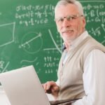 Professor sorridente com laptop nas mãos em sala de aula, com quadro verde ao fundo repleto de fórmulas matemáticas. Foto: Freepik