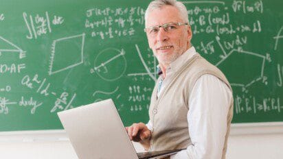 Professor sorridente com laptop nas mãos em sala de aula, com quadro verde ao fundo repleto de fórmulas matemáticas. Foto: Freepik
