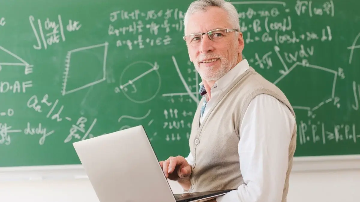 Professor sorridente com laptop nas mãos em sala de aula, com quadro verde ao fundo repleto de fórmulas matemáticas. Foto: Freepik