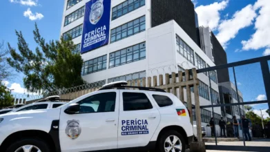 Viatura branca do concurso IGP RS estacionada em frente à sede do Instituto-Geral de Perícias do Rio Grande do Sul, com banner azul escrito "Perícia Criminal Rio Grande do Sul" fixado na fachada do prédio.