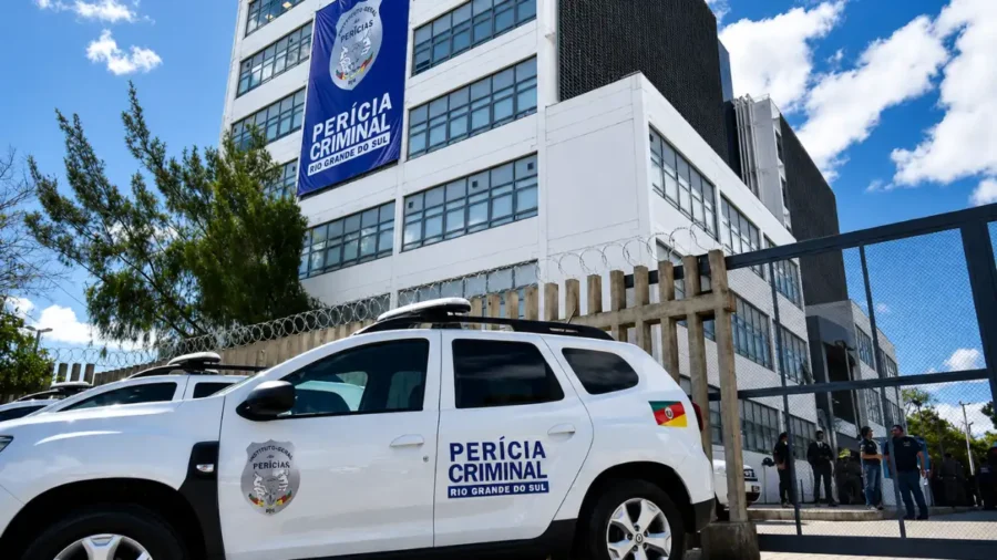 Viatura branca do concurso IGP RS estacionada em frente à sede do Instituto-Geral de Perícias do Rio Grande do Sul, com banner azul escrito "Perícia Criminal Rio Grande do Sul" fixado na fachada do prédio.