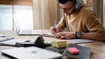 Homem usando fones de ouvido estuda em uma mesa com notebook, cadernos e blocos de anotações.
