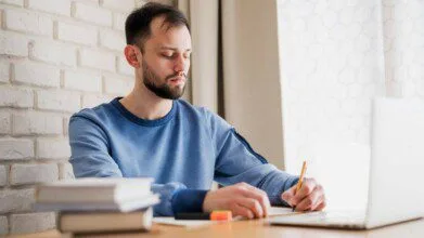 Homem sentado à mesa com caderno e lápis, concentrado nos estudos, ao lado de um notebook e pilha de livros, em ambiente claro e arejado.
