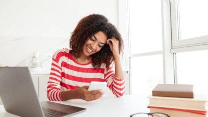 Mulher sorridente usando camiseta listrada vermelha e branca olha para o celular sentada à mesa com notebook, livros e óculos. Foto: drobotdean/Freepik