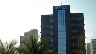Fachada do edifício sede do Ministério Público do Estado do Espírito Santo (MPES), um prédio vertical de vidro azul sob céu claro e palmeiras em primeiro plano.