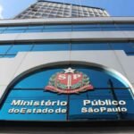 A imagem mostra a fachada de um edifício moderno com amplas janelas de vidro espelhado azul. Em destaque, na entrada do prédio, está o brasão do Estado de São Paulo com a sigla "SP" e os dizeres "Pro Brasilia Fiant Eximia". Abaixo do brasão, lê-se "Ministério Público do Estado de São Paulo" em letras metálicas. A foto é tirada de baixo para cima, mostrando a imponência da construção contra o céu azul.