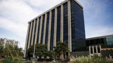 A imagem mostra o imponente prédio-sede da Prefeitura do Rio de Janeiro, com fachada moderna composta por grandes painéis de vidro escuro e colunas verticais claras que se estendem por toda a altura da edificação. O edifício possui diversos andares e está localizado em uma área arborizada com jardins e esculturas na frente. À esquerda, vê-se parte de outros prédios residenciais e, ao fundo, o céu azul com nuvens claras. A arquitetura transmite um ar institucional e oficial.