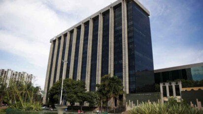 A imagem mostra o imponente prédio-sede da Prefeitura do Rio de Janeiro, com fachada moderna composta por grandes painéis de vidro escuro e colunas verticais claras que se estendem por toda a altura da edificação. O edifício possui diversos andares e está localizado em uma área arborizada com jardins e esculturas na frente. À esquerda, vê-se parte de outros prédios residenciais e, ao fundo, o céu azul com nuvens claras. A arquitetura transmite um ar institucional e oficial.