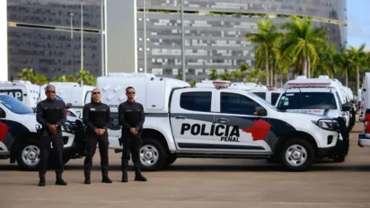 Três agentes da Polícia Penal de Minas Gerais uniformizados em frente a viaturas brancas com o logotipo da corporação estacionadas em área urbana. Foto: Divulgação/PPMG