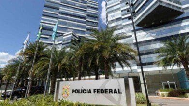 A imagem mostra a fachada moderna do edifício-sede da Polícia Federal do Brasil, com design arquitetônico contemporâneo, composto por estruturas espelhadas em tons de cinza e preto. Em frente ao prédio, há palmeiras alinhadas e uma placa com o brasão da instituição e os dizeres “POLÍCIA FEDERAL”. Três mastros com bandeiras hasteadas também estão presentes, juntamente com um veículo oficial estacionado à esquerda. O céu está azul com algumas nuvens.