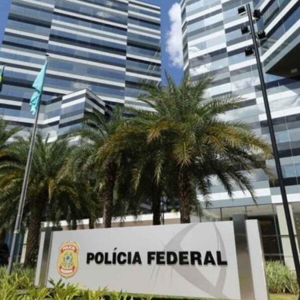 A imagem mostra a fachada moderna do edifício-sede da Polícia Federal do Brasil, com design arquitetônico contemporâneo, composto por estruturas espelhadas em tons de cinza e preto. Em frente ao prédio, há palmeiras alinhadas e uma placa com o brasão da instituição e os dizeres “POLÍCIA FEDERAL”. Três mastros com bandeiras hasteadas também estão presentes, juntamente com um veículo oficial estacionado à esquerda. O céu está azul com algumas nuvens.