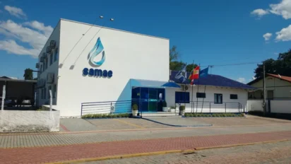 Fachada da sede do concurso SAMAE Timbó SC com logotipo azul e bandeiras ao lado