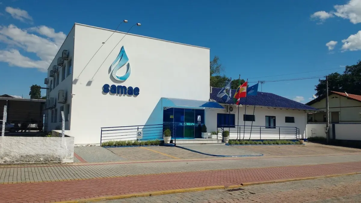 Fachada da sede do concurso SAMAE Timbó SC com logotipo azul e bandeiras ao lado
