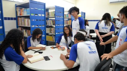 Grupo de estudantes em biblioteca escolar, usando uniformes da rede estadual de Sergipe enquanto leem, escrevem e conversam ao redor de mesas. Foto: Divulgação/Maria Odília/SEDUC-SE