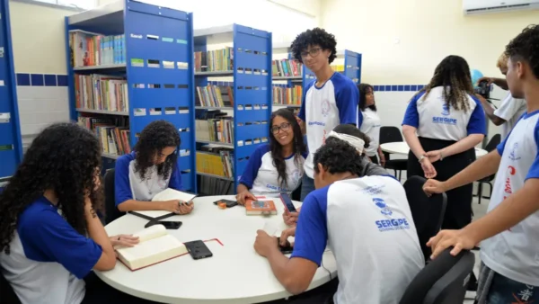 Concurso SEDUC/SE abre 104 vagas imediatas de até R$ 5,1 mil 2 Grupo de estudantes em biblioteca escolar, usando uniformes da rede estadual de Sergipe enquanto leem, escrevem e conversam ao redor de mesas. Foto: Divulgação/Maria Odília/SEDUC-SE
