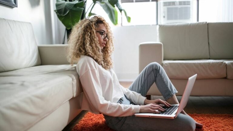 Mulher sentada no chão entre dois sofás, usando notebook no colo em ambiente iluminado com tapete laranja e planta ao fundo. Foto: Thought Catalog/Unsplash