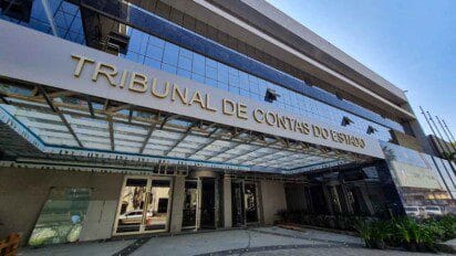 Fachada espelhada do prédio do Tribunal de Contas do Estado com letreiro dourado e cobertura de vidro na entrada.