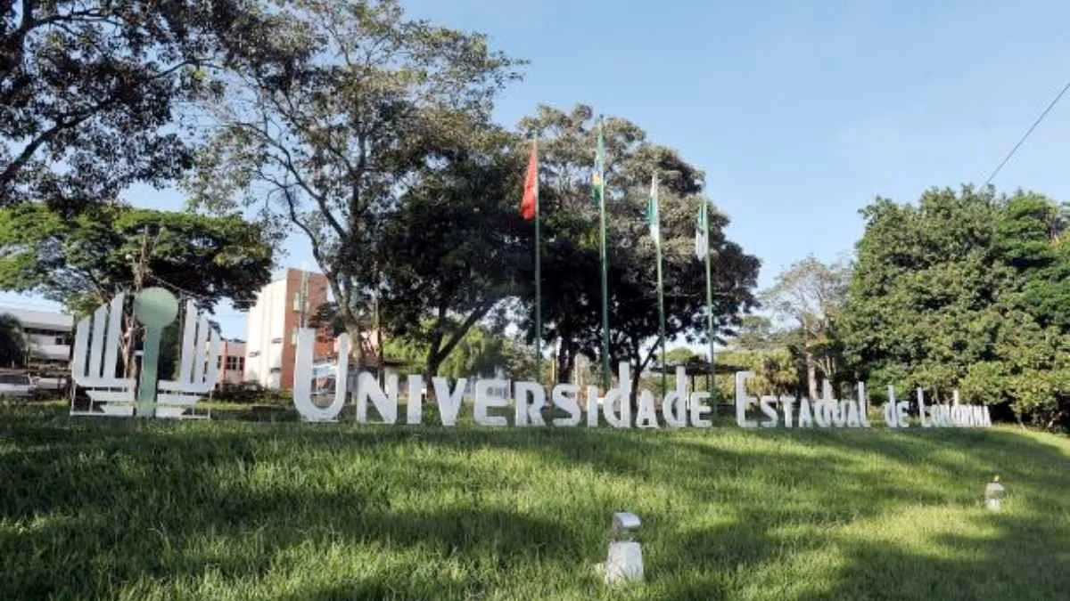 Vista da entrada principal da Universidade Estadual de Londrina (UEL), no Paraná, com o letreiro institucional em destaque sobre o gramado. Ao fundo, aparecem bandeiras hasteadas e prédios acadêmicos cercados por árvores, compondo um cenário arborizado e bem cuidado. A imagem foi registrada em um dia ensolarado, com céu azul e iluminação natural, evidenciando a infraestrutura e o ambiente do campus universitário.