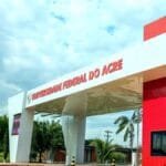 Entrada principal da Universidade Federal do Acre com estrutura moderna nas cores branca e vermelha, rodeada por palmeiras e céu parcialmente nublado ao fundo