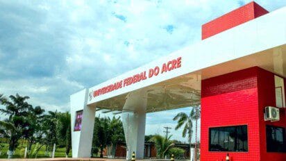 Entrada principal da Universidade Federal do Acre com estrutura moderna nas cores branca e vermelha, rodeada por palmeiras e céu parcialmente nublado ao fundo