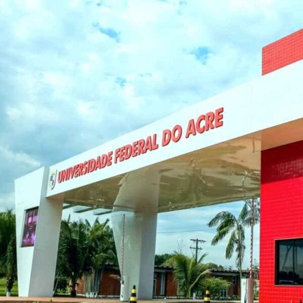 Entrada principal da Universidade Federal do Acre com estrutura moderna nas cores branca e vermelha, rodeada por palmeiras e céu parcialmente nublado ao fundo