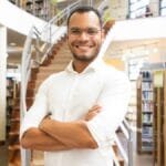 UNEAL - Homem jovem, com barba rala e óculos de grau, aparece sorrindo com os braços cruzados, vestindo camisa branca de manga longa. Ele está em uma biblioteca moderna e iluminada, com estantes cheias de livros ao fundo e uma escada em espiral ao centro. A atmosfera é acolhedora e acadêmica, transmitindo um ambiente de estudo, ensino ou trabalho intelectual.