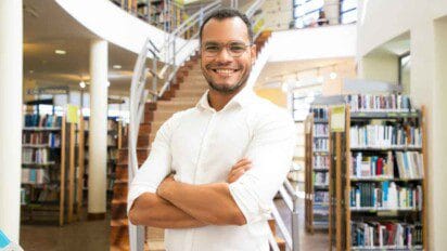 UNEAL - Homem jovem, com barba rala e óculos de grau, aparece sorrindo com os braços cruzados, vestindo camisa branca de manga longa. Ele está em uma biblioteca moderna e iluminada, com estantes cheias de livros ao fundo e uma escada em espiral ao centro. A atmosfera é acolhedora e acadêmica, transmitindo um ambiente de estudo, ensino ou trabalho intelectual.