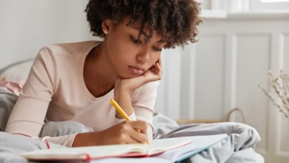 Mulher deitada em uma cama escrevendo em um caderno com expressão concentrada. Foto: wayhomestudio/Freepik