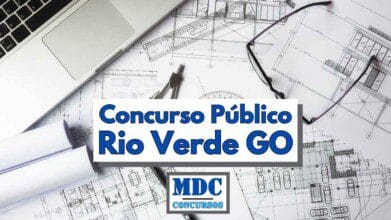 Imagem ilustrativa de um concurso público em Rio Verde, Goiás. No centro da imagem há uma faixa com os dizeres "Concurso Público Rio Verde GO" em letras azuis destacadas. Ao fundo, estão dispostos elementos relacionados à arquitetura e engenharia, como projetos técnicos, óculos, compasso, esquadros e parte de um notebook prateado aberto sobre a mesa