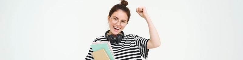 IFG abre 250 vagas em pós-graduação MBA gratuito 100% EaD 2 Mulher sorridente com óculos e fones no pescoço comemora com o punho erguido enquanto segura cadernos.