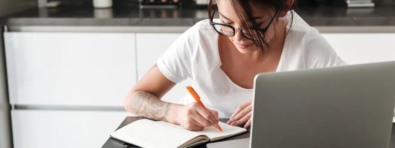 Jarinu/SP lança concurso com salários de até R$ 13.455,00 2 Mulher jovem escrevendo em um caderno à mesa, com um notebook aberto à frente, em ambiente interno.