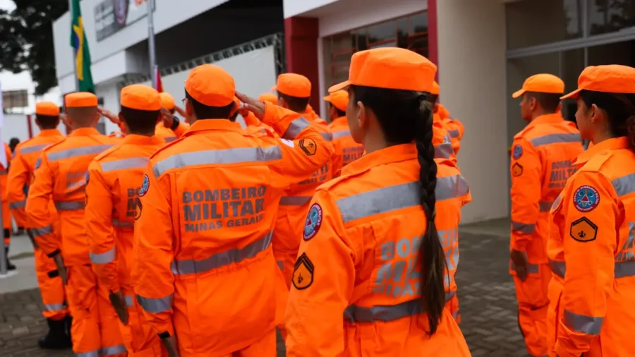 Fileira de bombeiros militares de Minas Gerais (Corpo de Bombeiros) em formação, vestindo uniformes operacionais na cor laranja com faixas refletivas e bonés, prestando continência em ambiente externo.