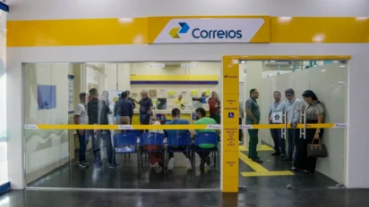 Interior de uma agência dos Correios com fachada de vidro, logomarca amarela e azul, e pessoas aguardando atendimento em cadeiras azuis ou em pé próximo aos guichês.