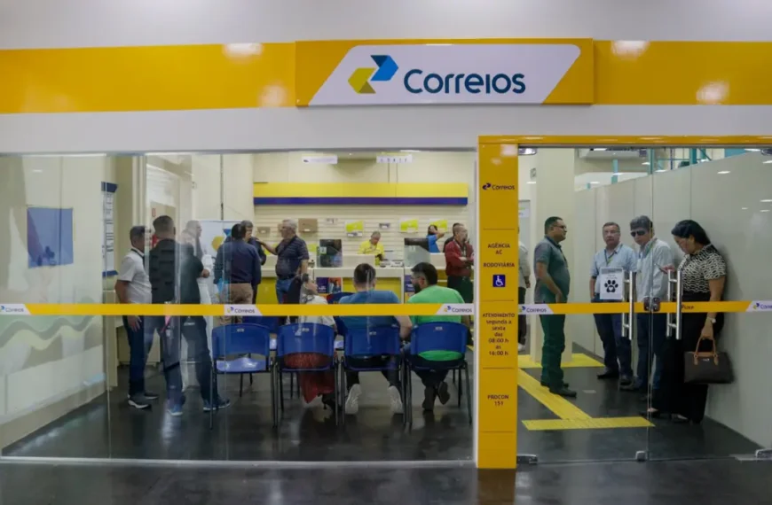 Interior de uma agência dos Correios com fachada de vidro, logomarca amarela e azul, e pessoas aguardando atendimento em cadeiras azuis ou em pé próximo aos guichês.