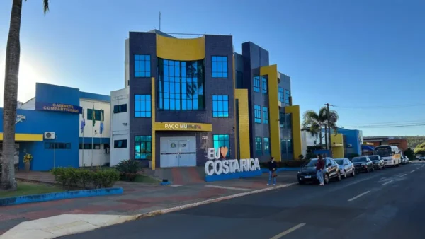 Fachada do Paço Municipal de Costa Rica, Mato Grosso do Sul, com arquitetura em tons de azul e amarelo, letreiro com o nome da cidade à frente e carros estacionados na rua lateral.
