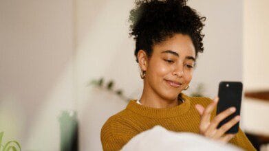 Processos Seletivos 7 Mulher sorrindo e olhando para a tela do celular enquanto está sentada em ambiente interno.