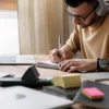 Homem usando fones de ouvido escreve em um caderno ao lado de um notebook em uma mesa com materiais de estudo. Foto: Freepik