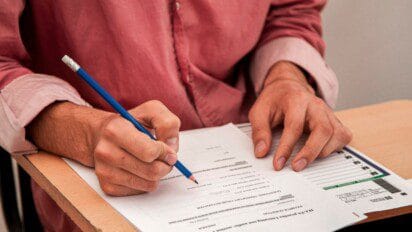 Pessoa preenchendo prova ou formulário com lápis em carteira escolar. Foto: azerbaijan_stockers/Freepik