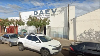 Fachada de um prédio branco com a inscrição "DAEV - Departamento de Água e Esgotos de Valparaíso". Há carros estacionados na frente e árvores de pequeno porte na calçada sob um céu com nuvens.
